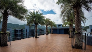 Een houten terras met palmbomen, een glazen balustrade en uitzicht op gebouwen en bergen.