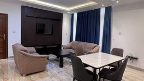 Ein modernes Wohnzimmer mit einer Sofagarnitur, einem an einer Holzplatte montierten Fernseher, einem Esstisch mit Stühlen und einem großen Fenster mit Vorhängen.