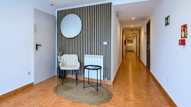 Een gang met een radiator, een stoel, een klein tafeltje en een ronde spiegel aan de muur.