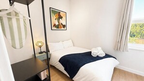 A neatly made bed with white linens and a dark blue blanket, a hanging garment, a framed abstract painting, and a window with curtains. - Exclusive Studio in the Heart of Hounslow! (Greater London)