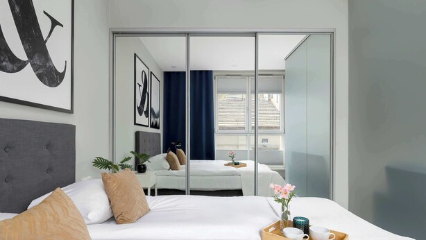 Ein modernes Hotelzimmer mit einem großen Bett, einem Nachttisch mit Tablett, einem Spiegel und Blick durch eine Glastür.
