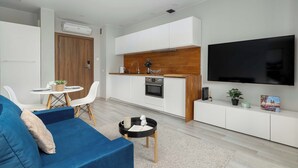 Ein modernes Wohnzimmer mit einem blauen Sofa, einem kleinen runden Tisch und einer Küchenzeile mit weißen Schränken und einem schwarzen Fernseher.