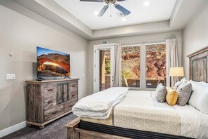 A bedroom with a bed, a TV, a wooden dresser, and a view of a rocky landscape through the window. - Coral Ridge 2076, Near Golf Course, Community Amenities! (Washington)