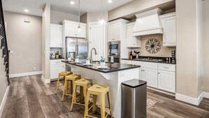 Una cocina moderna con armarios blancos, electrodomésticos de acero inoxidable, una isla central con taburetes y un elemento decorativo en la pared.