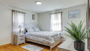 Ein Schlafzimmer mit Bett, Nachttisch, Lampe, Vorhängen, einem gerahmten Bild und einer Topfpflanze.