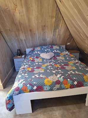 A bedroom with a wooden sloped ceiling, a floral-patterned bedspread, a bedside table with a lamp, and a small drawer unit. - Renovated to make your stay "just like home". Pets allowed! (Nonards)