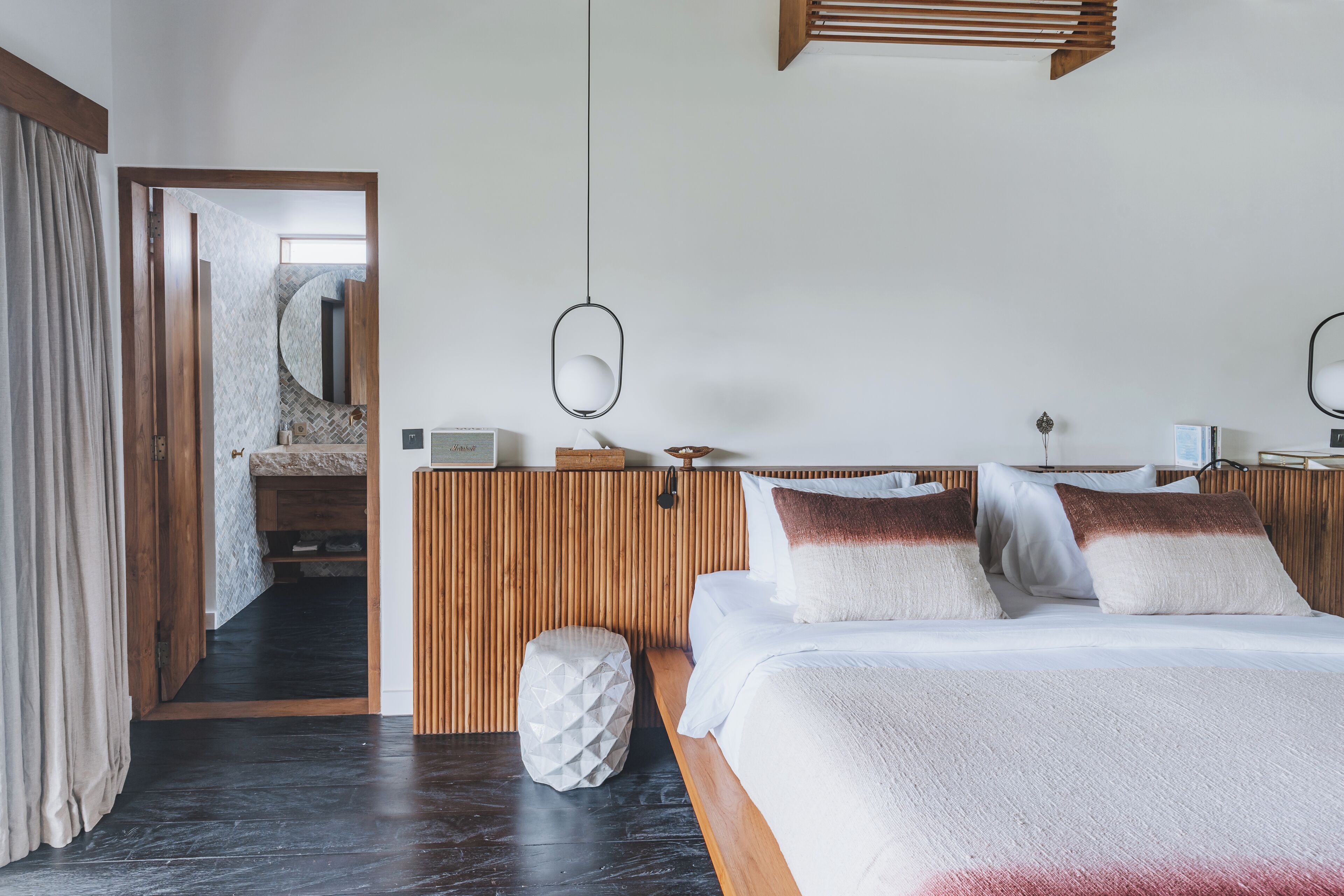 A modern hotel room with a wooden headboard, a large bed with white bedding, a bedside table with a lamp, and a view of the bathroom through an open door.