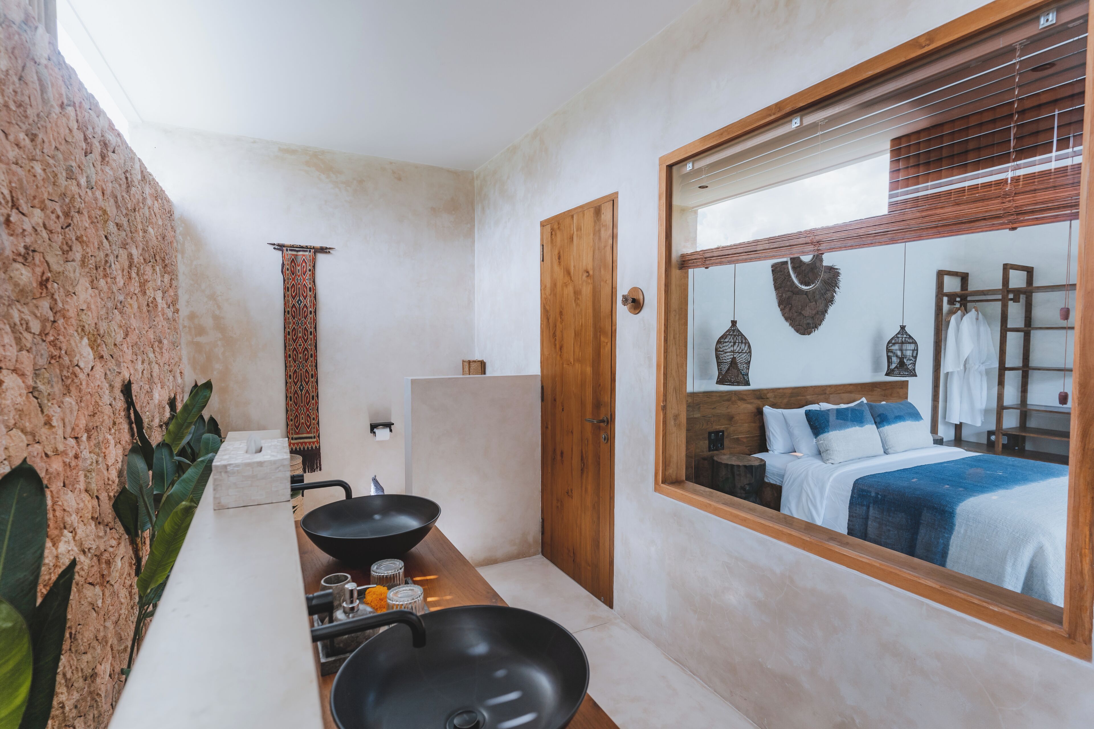 A bathroom with a stone wall, a wooden door, a large mirror, a bed with a blue and white bedspread, and a black sink.