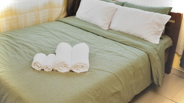 A bed with green bedding, white pillows, and rolled towels.