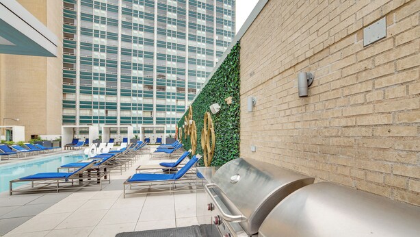 Un espace piscine aménagé sur le toit, avec des chaises longues, un barbecue et une vue sur un immeuble de grande hauteur.