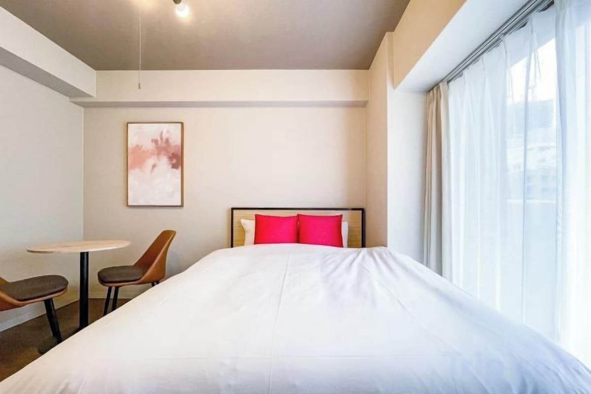 A hotel room with a large bed, a small table, two chairs, a painting on the wall, and a window with curtains.