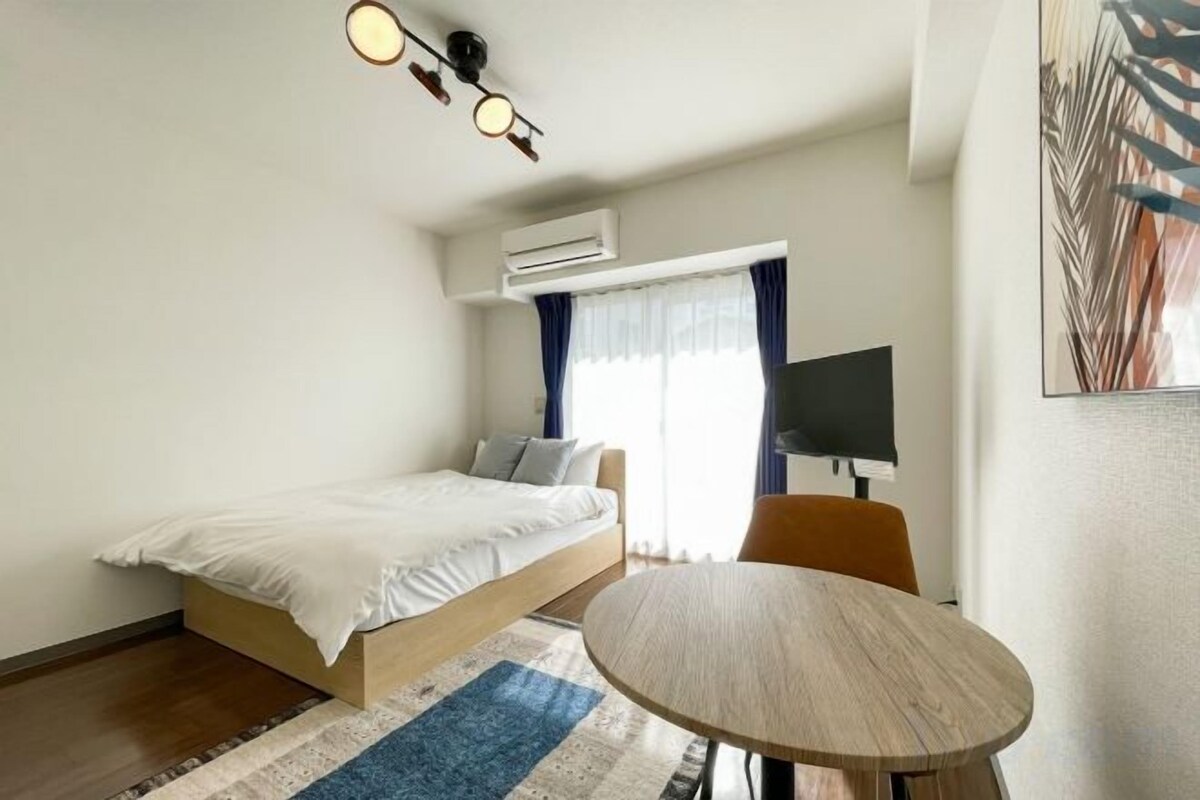 A hotel room with a bed, a round table, a TV, and an air conditioning unit.