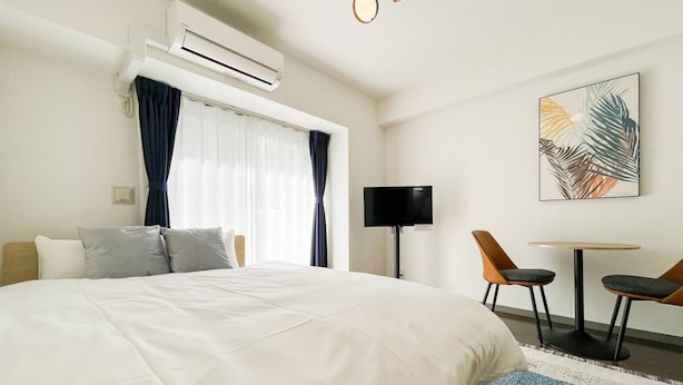 Quarto de hotel com uma cama, televisão, escrivaninha e duas cadeiras, e um ar condicionado.