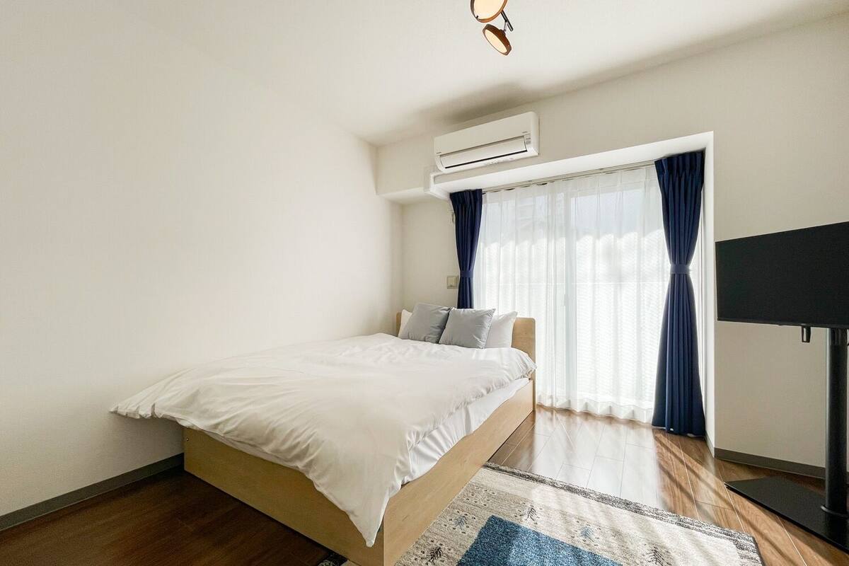 A bedroom with a bed, an air conditioning unit, a television, and a window with curtains.