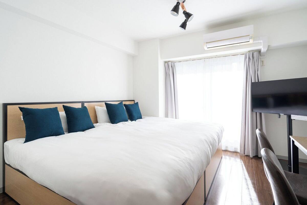 A hotel room with a large bed, a flat-screen TV, an air conditioning unit, and a window with curtains.