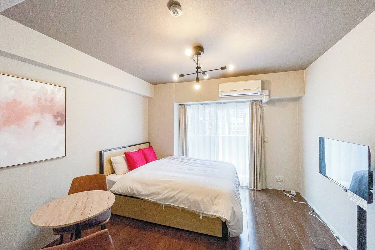 A hotel room with a bed, a small table, a chair, an air conditioning unit, and a television.