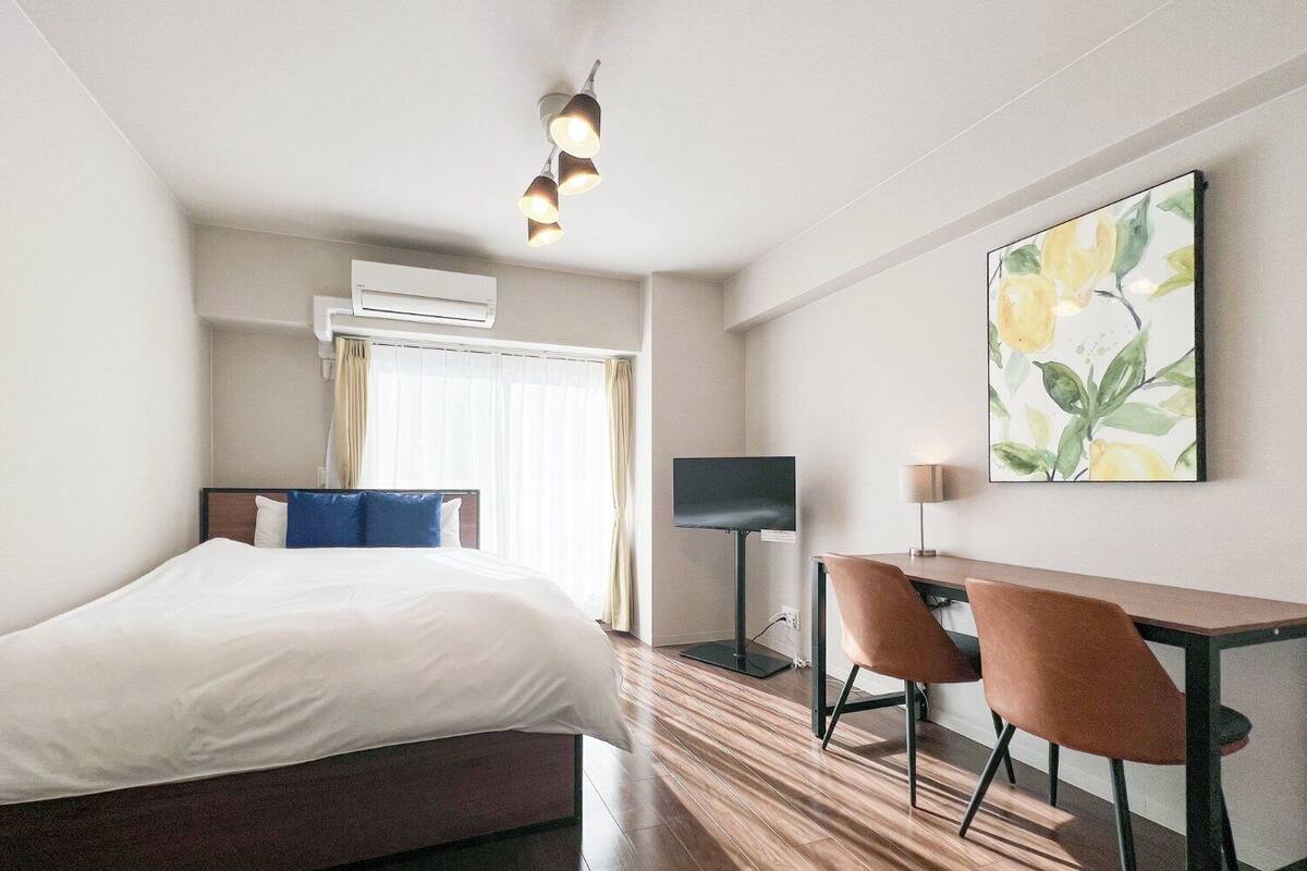 A hotel room with a bed, a desk, two chairs, a flat-screen TV, an air conditioning unit, and a painting on the wall.