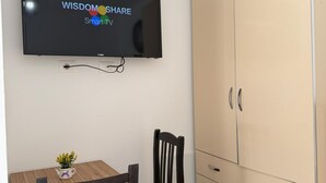 Quarto com televisão fixada na parede exibindo 'WISDOMOS SHARE Smart TV', uma mesa de madeira com uma cadeira e um guarda-roupa bege espaçoso com gavetas.