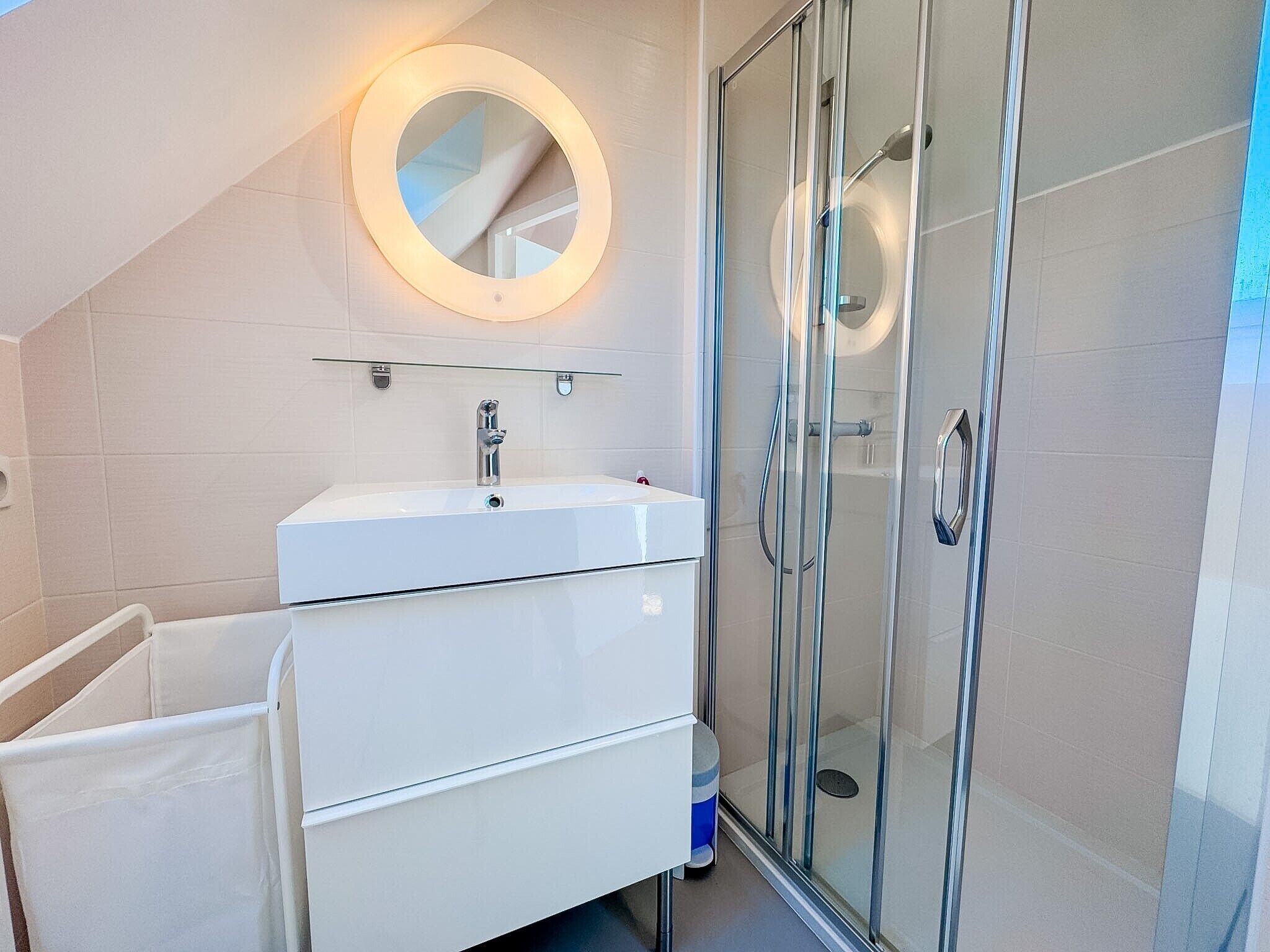 A modern bathroom with a glass shower enclosure, a white sink, and a round mirror with a light.