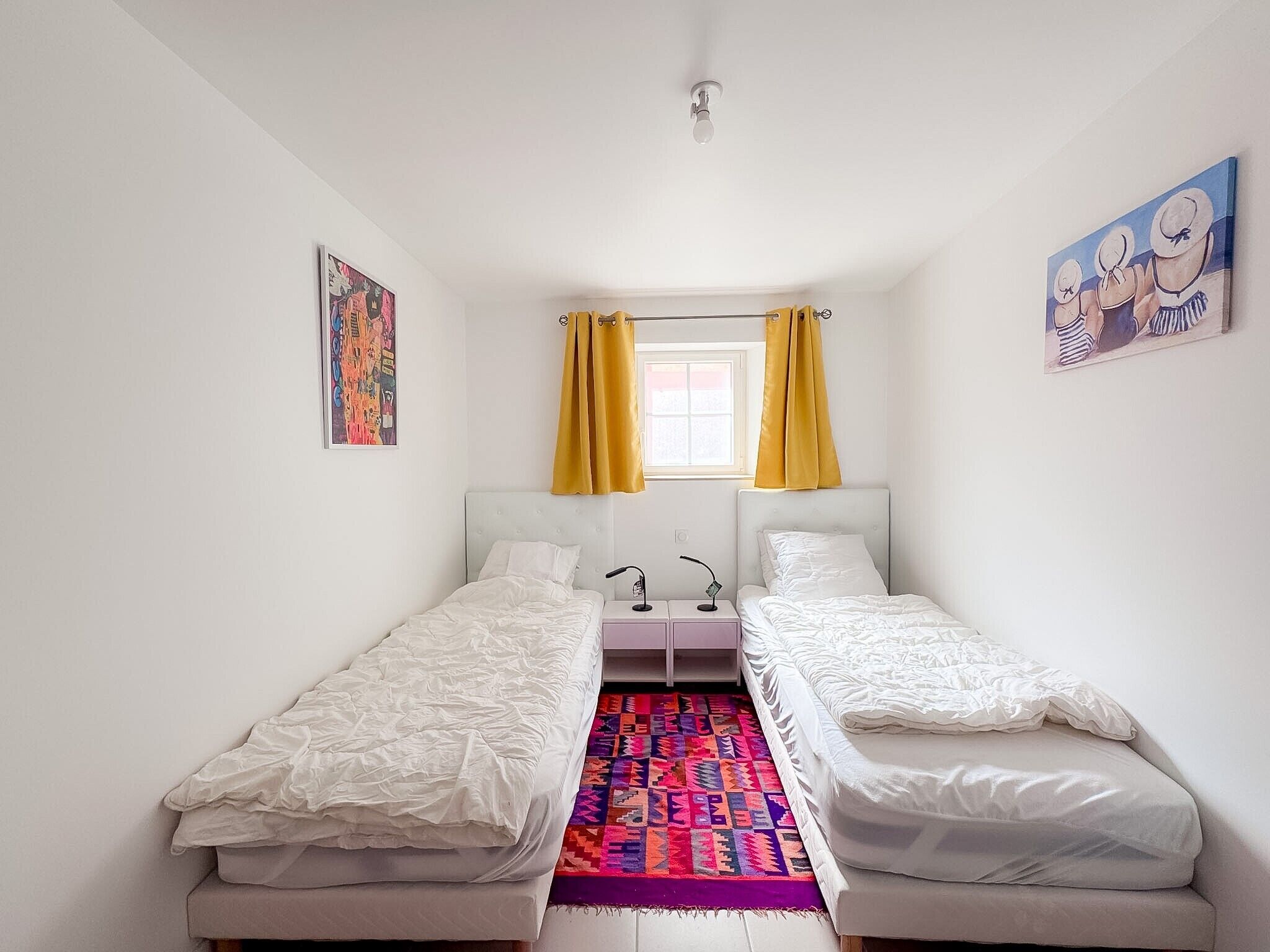 A small, bright room with two single beds, a colorful rug, and a window with yellow curtains.