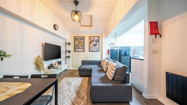 A modern living room with a flat-screen TV, a grey sofa, a dining table with chairs, and a large window. - Apartment in Cite Center (Manchester)