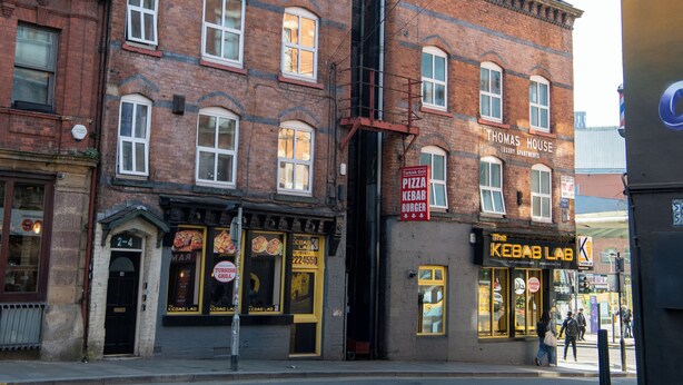 A brick building with multiple windows, a sign for 'Thomas House' indicating it is a luxury apartment building, and a 'Kebab Lab' restaurant with a visible menu. - Apartment in Cite Center (Manchester)