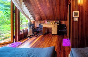 Une pièce aux murs en bois, avec un bureau, deux chaises, un ventilateur et une fenêtre donnant sur un paysage verdoyant.
