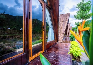 Une porte vitrée à ossature en bois donne accès à un jardin agrémenté d’un bassin, entouré de plantes tropicales, avec une chaîne de montagnes en toile de fond.