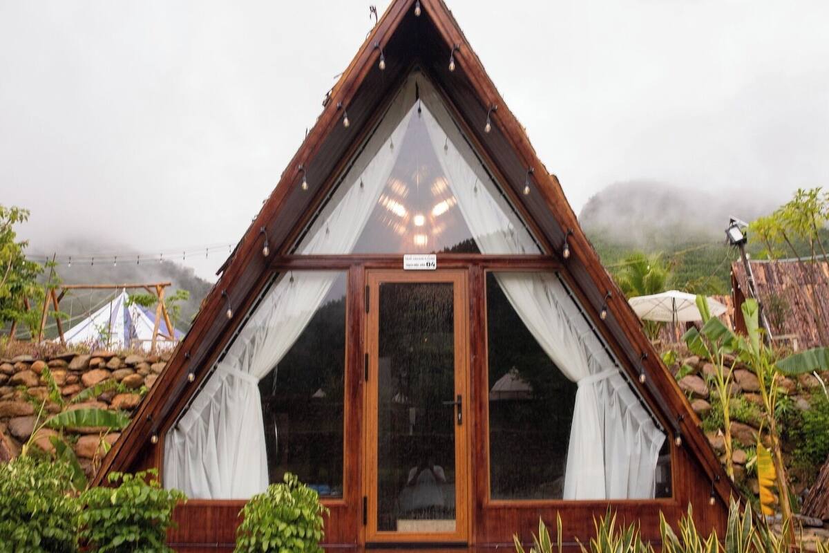 A triangular wooden structure with a large window and a door, surrounded by plants.