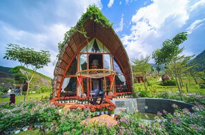 A thatched-roof structure with a garden in front, surrounded by greenery. - LEAF VILLAGE & FARM LÀNG LÁ (Da Nang)