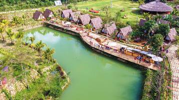 A riverside resort with thatched-roof huts, a wooden walkway, and a calm river.