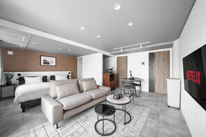 Moderni hotellihuone, jossa on sänky, sohva, kaksi pientä pöytää, televisio ja minibaari.