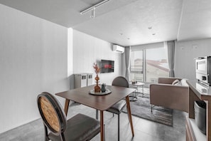 Quarto de hotel moderno com área de refeições, sofá, televisão e uma janela grande.