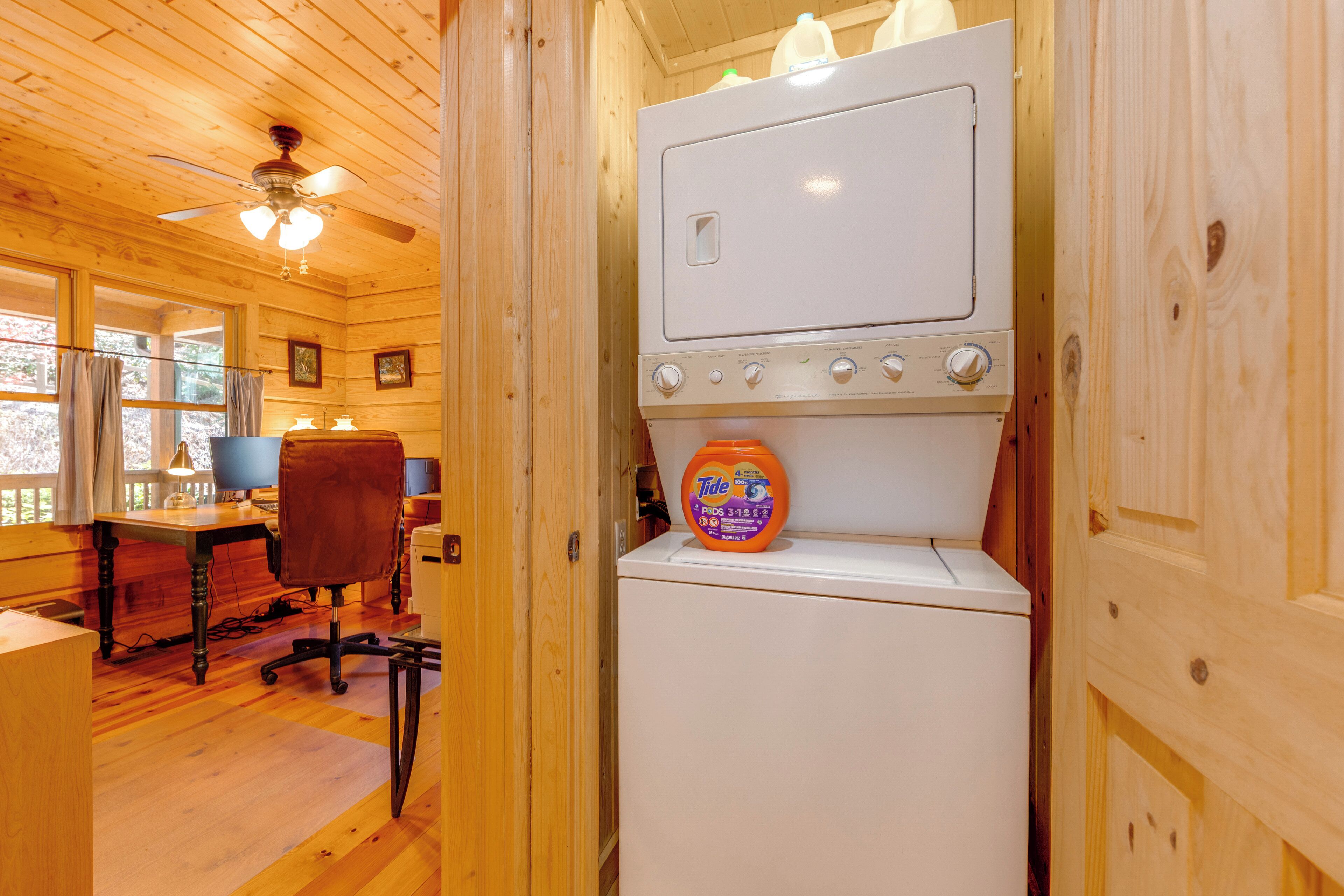 A compact room with a wooden interior, a white appliance with a detergent bottle, a ceiling fan, and a desk with a chair.