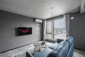 Un salon moderne avec un mur gris, un téléviseur fixé au mur affichant le logo « NETFLIX », une grande fenêtre avec des rideaux, un canapé bleu et une petite table ronde sur laquelle se trouvent une bouteille et un bol.