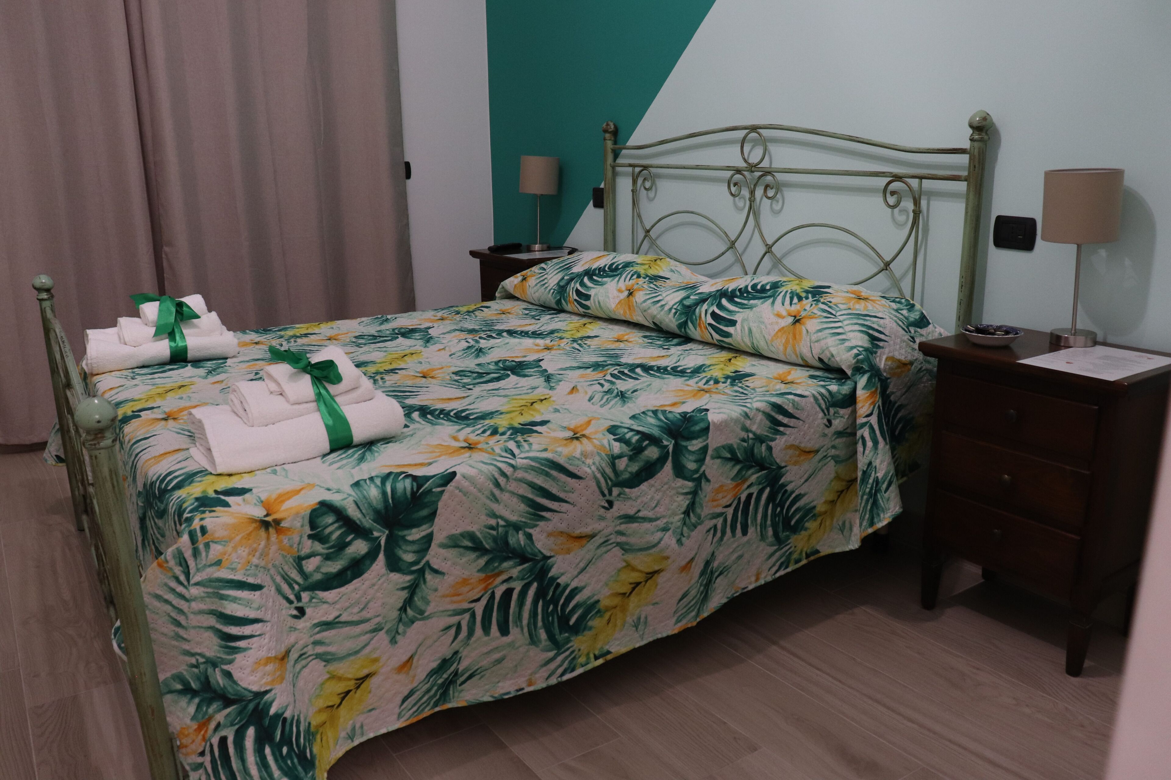 Una cama bien hecha con una colcha estampada, un par de toallas con un lazo verde, una mesita de noche con una lámpara y otra mesita de noche con una lámpara.