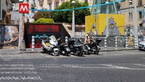 Calle con scooters estacionados, un letrero rojo con la letra 'M' y un edificio con techo a dos aguas.