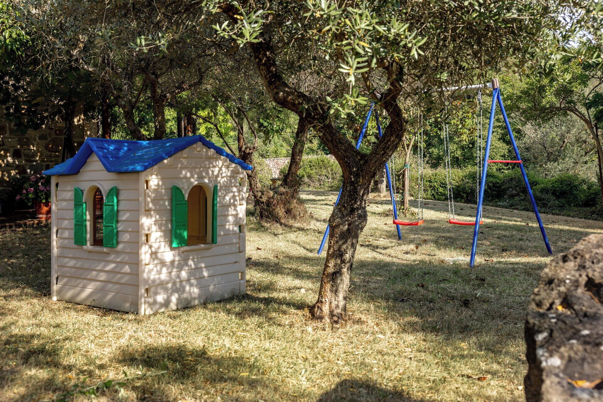 Ein kleines Spielhaus mit blauem Dach und grünen Fensterläden, eine Schaukel und ein Baum in einem Park.