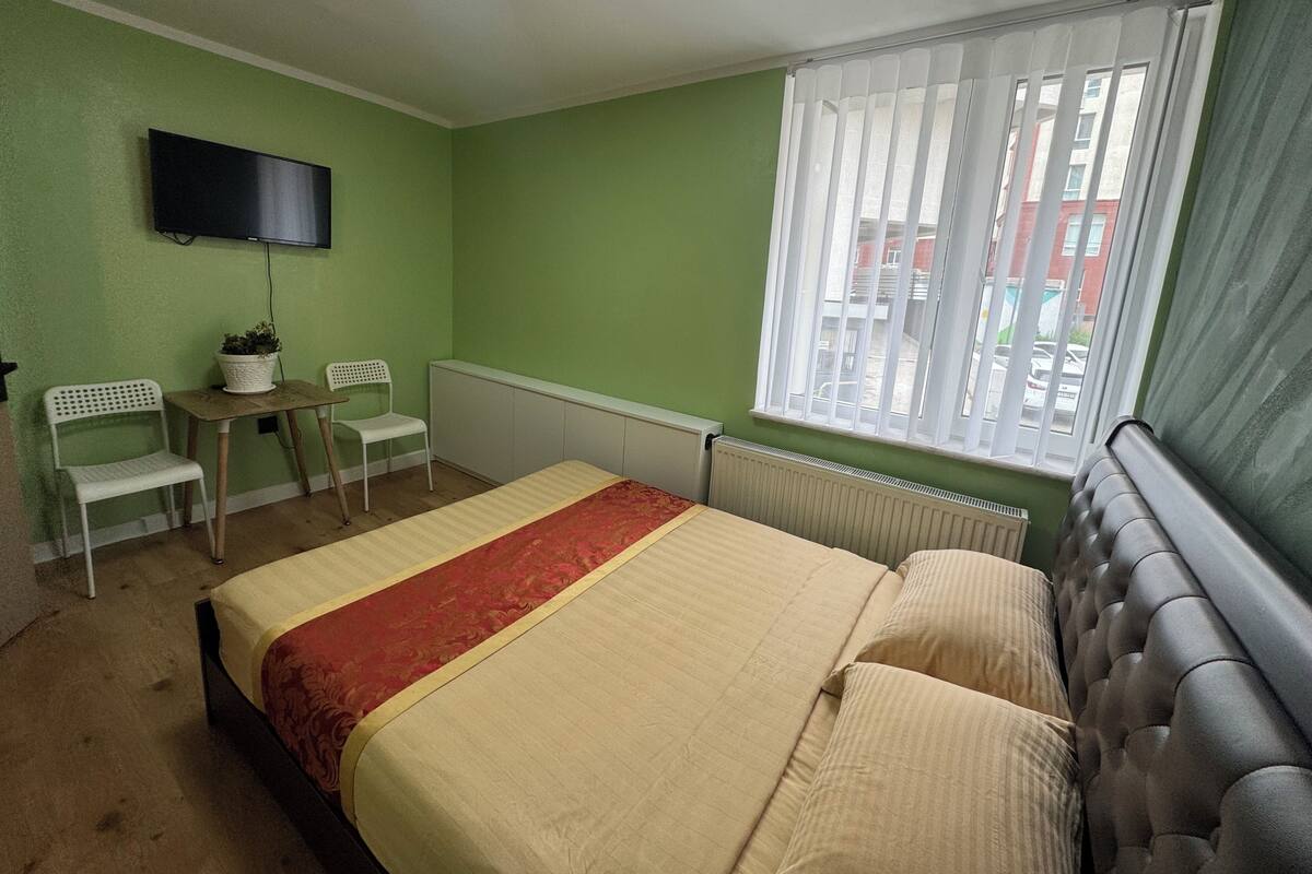A bedroom with a bed, a television, a window with blinds, and a radiator.