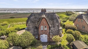 A thatched-roof house with a brick base, surrounded by greenery and a view of the sea.