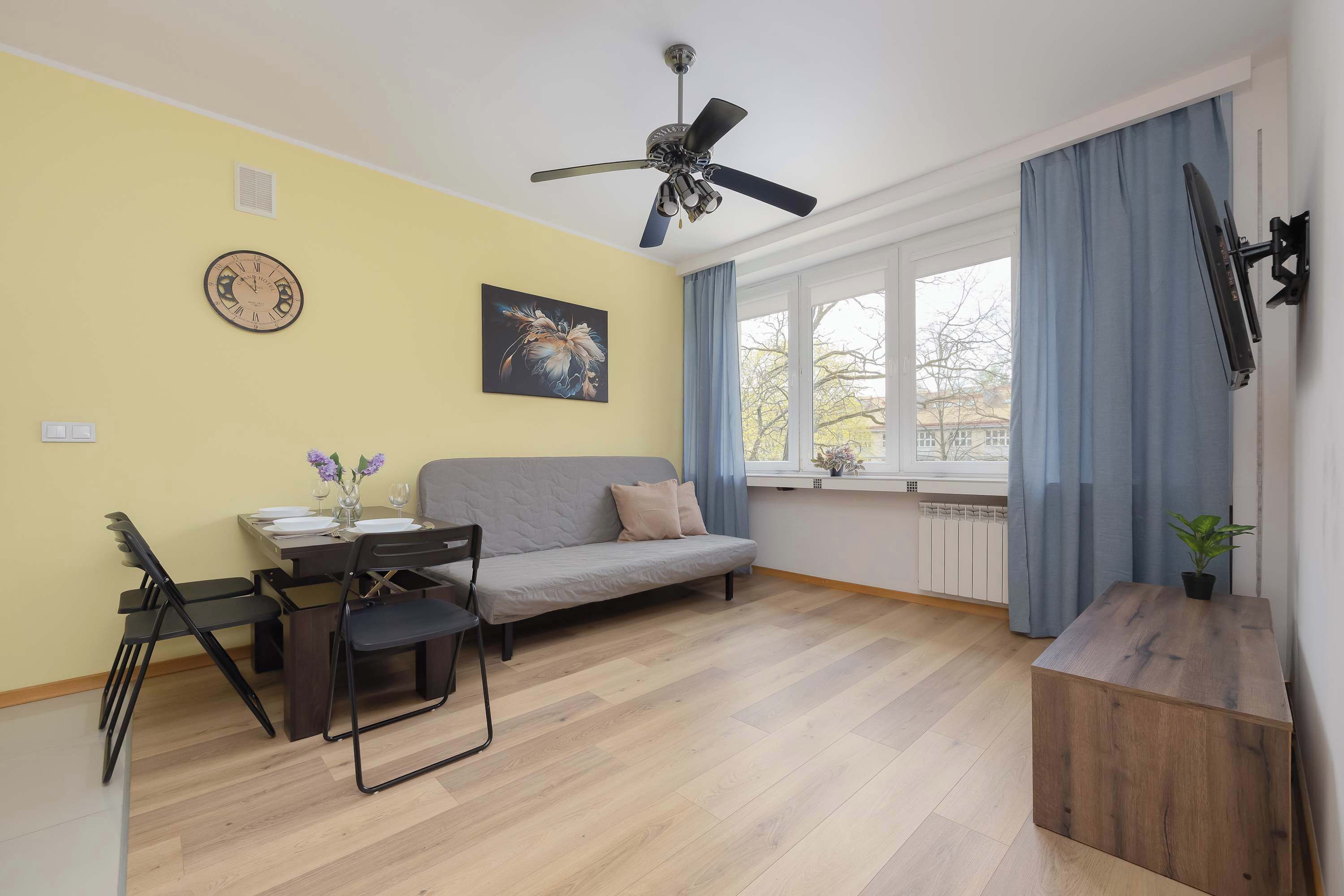 A room with a ceiling fan, a sofa, a dining table with chairs, a clock, a window with curtains, and a TV mounted on the wall.