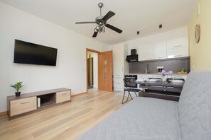 A modern living room with a flat-screen TV, wooden flooring, a ceiling fan, a sofa, a kitchen area with cabinets, and a dining table with chairs. - Cozy Apartment | Place to Work | Pet Friendly (Warszawa)
