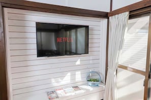 Una habitación con una televisión montada que muestra 'NETFLIX', una mesita con revistas y una ventana con cortinas.