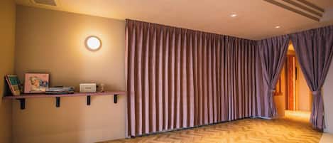 Habitación de hotel con suelo de madera tipo tablero de ajedrez, un ventanal con cortinas, una estantería con libros y una lámpara, y una puerta que da a otra habitación.