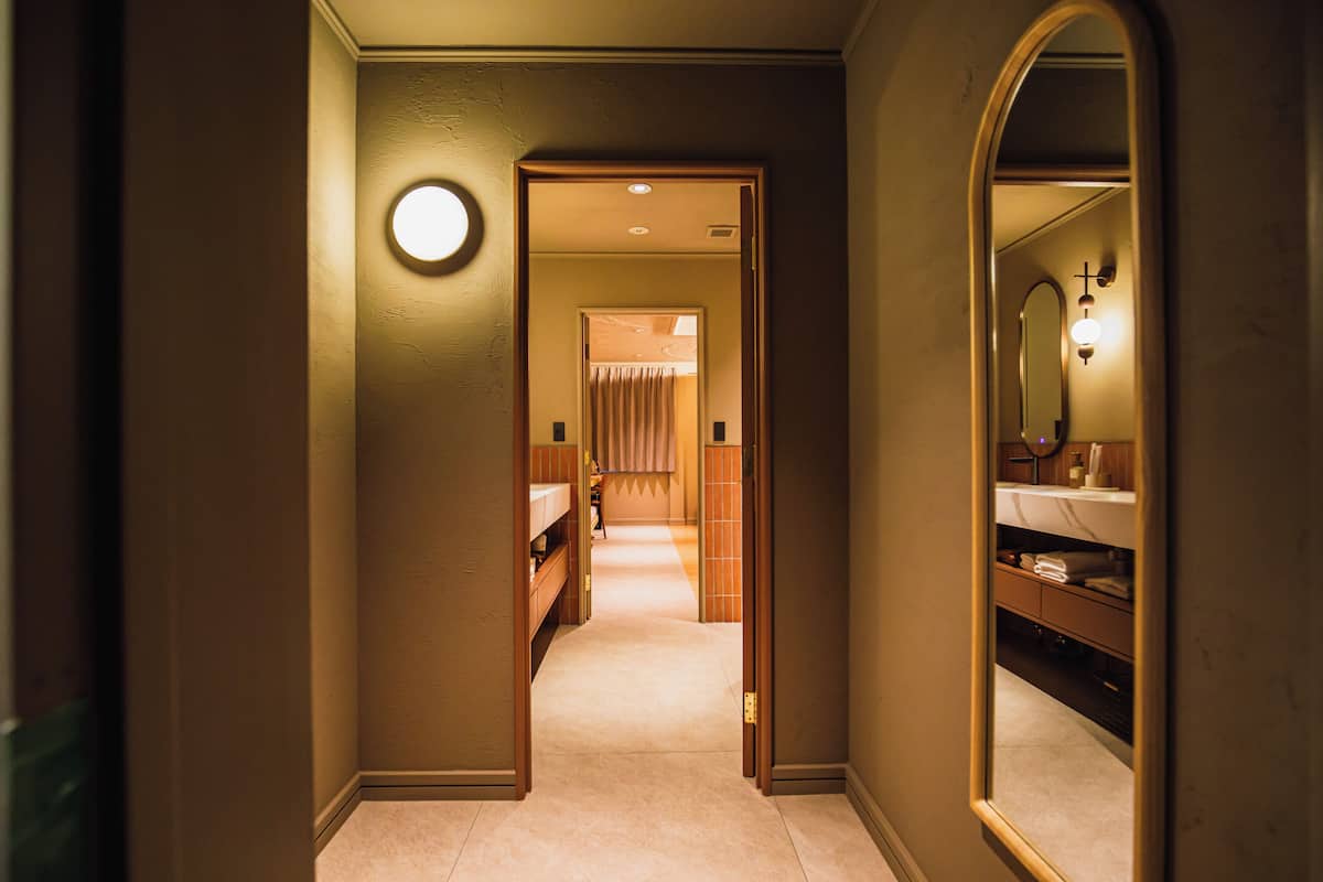 Pasillo de hotel con una lámpara redonda, un espejo y una puerta que da a una habitación con cama y baño.
