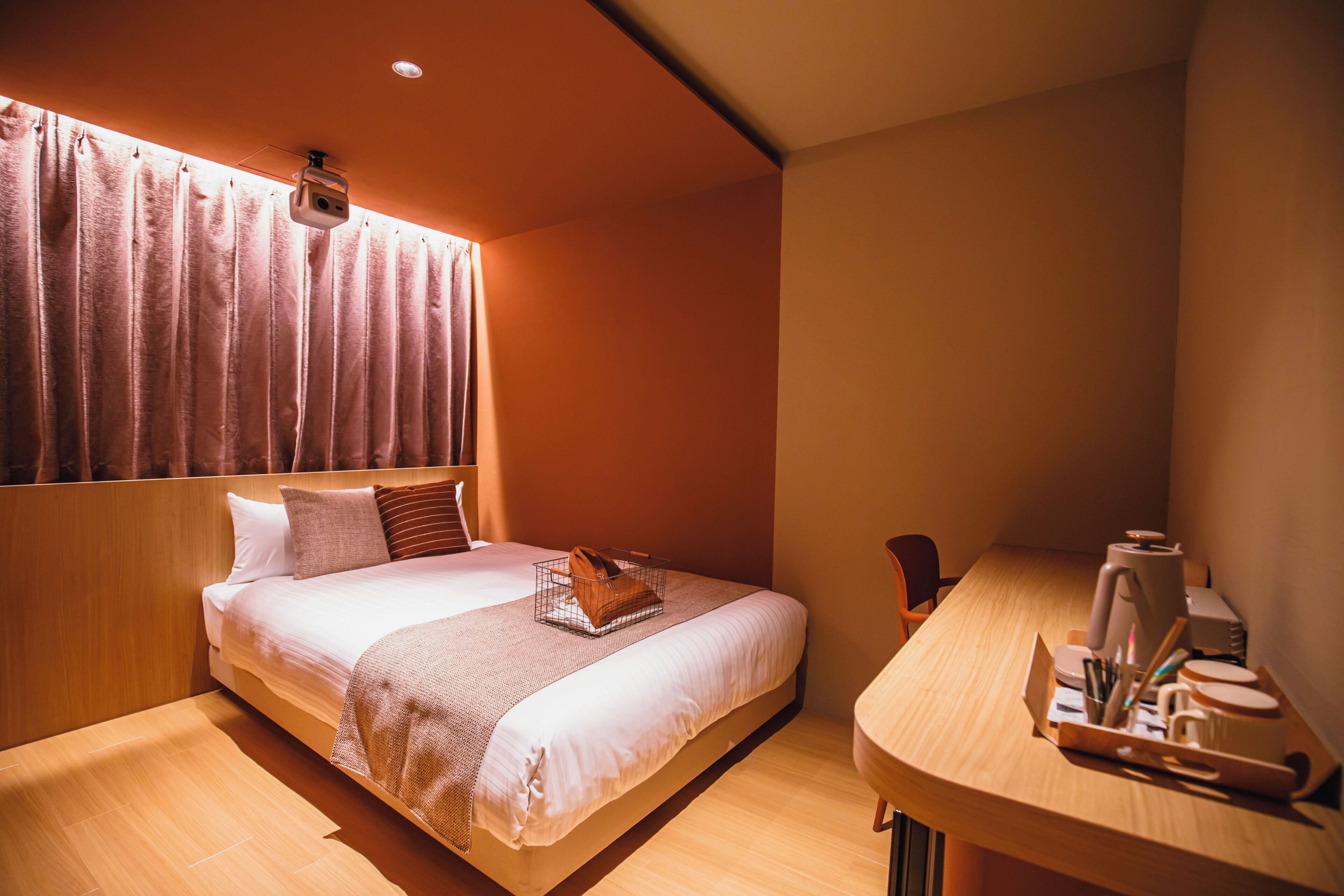 A neatly made bed with a headboard, a bedside table with a coffee maker, and a window with curtains.