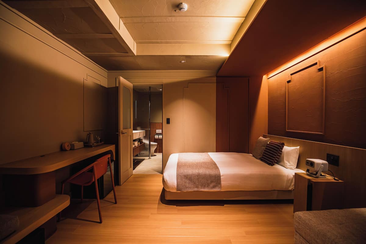 Una habitación de hotel moderna con cama, escritorio, silla y una mesita con lámpara.