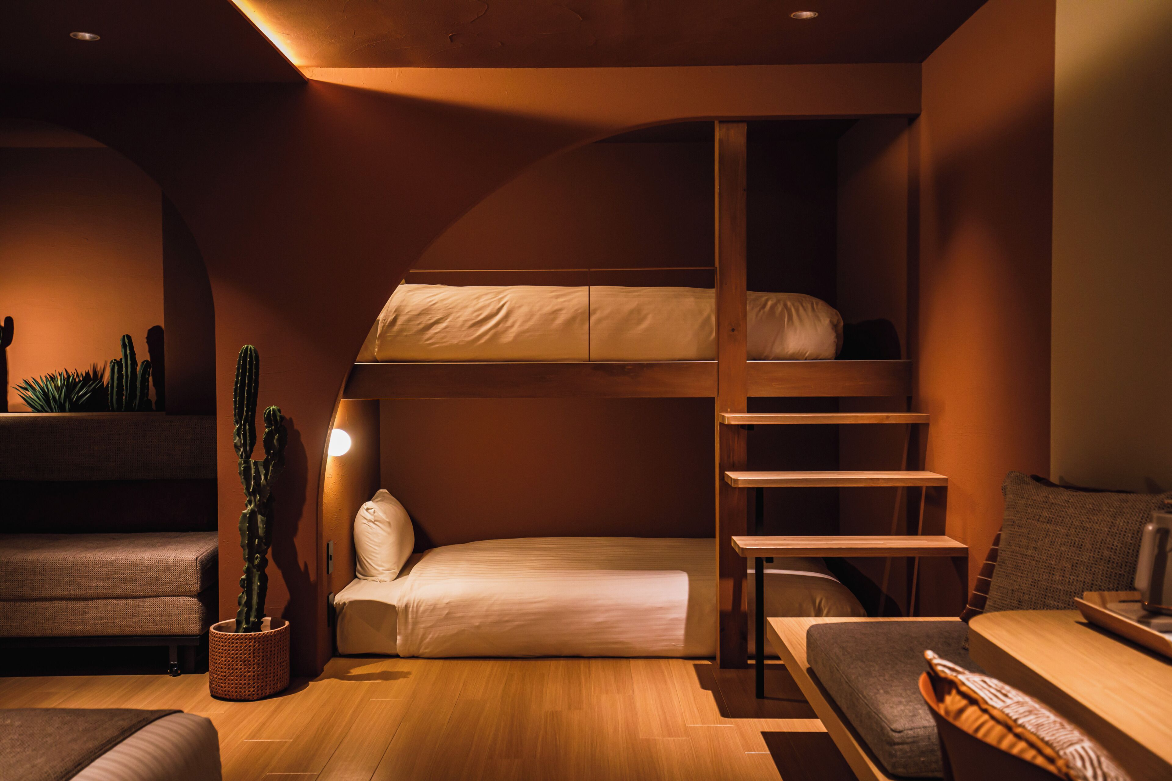 Una cama litera con escalera de madera, una mesita de noche pequeña, una silla, un sofá y una planta en maceta.