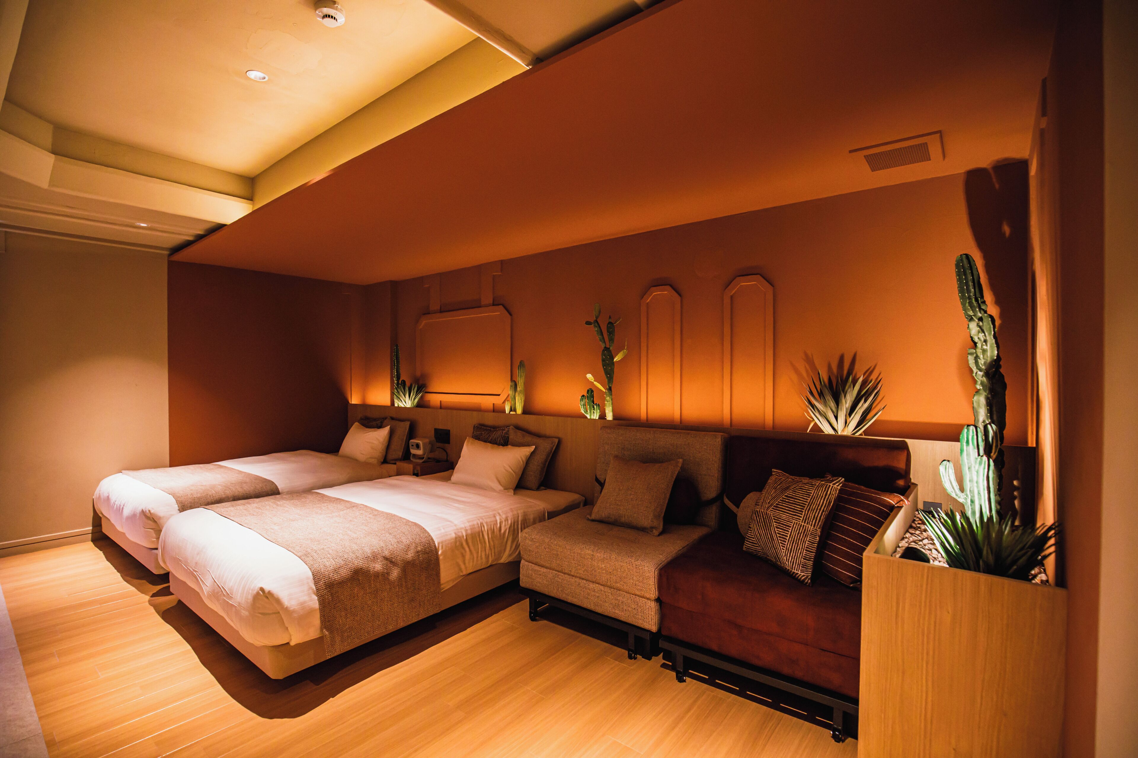 A hotel room with two beds, a sofa, a wooden floor, and a cactus plant.
