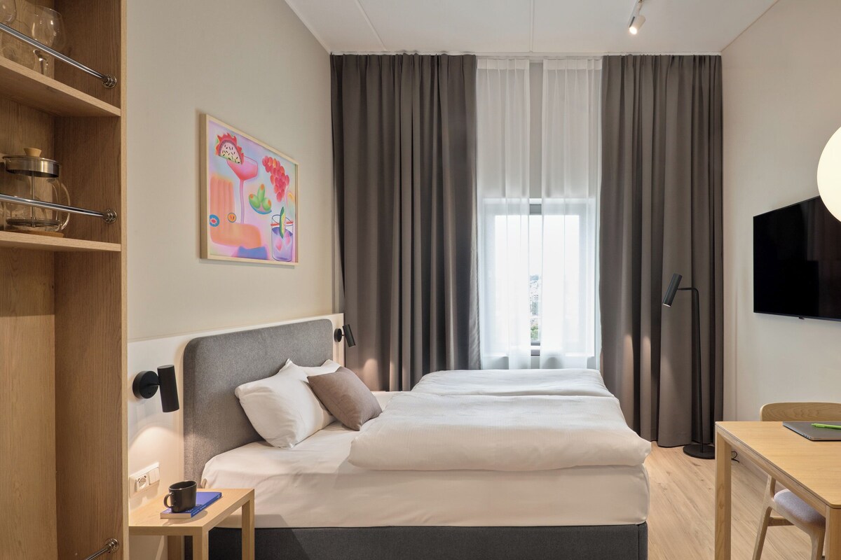 A hotel room with a bed, bedside table, desk, chair, TV, and a window with curtains.